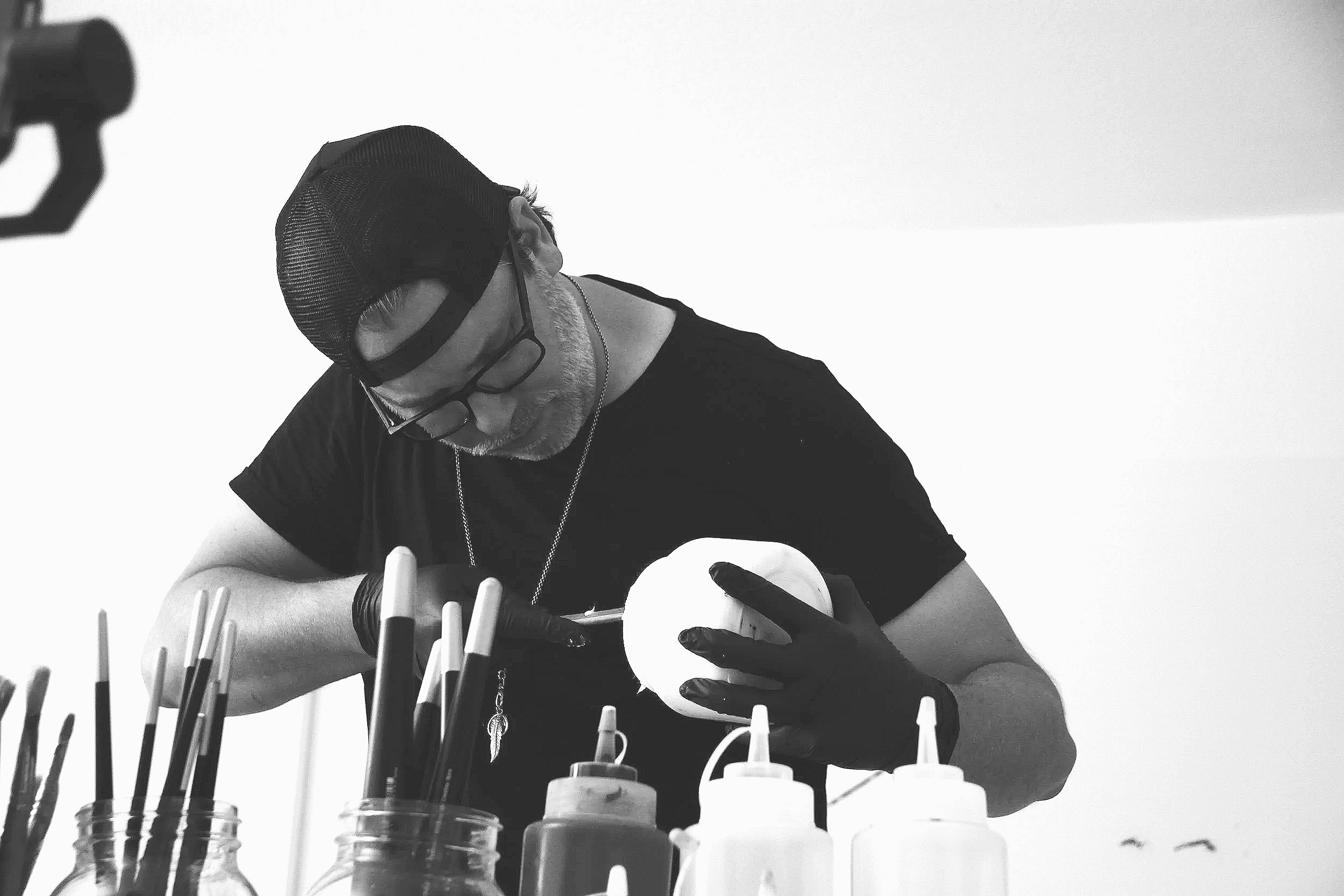 A black-and-white photograph of abstract artist Christopher Durst working in his Austin, Texas studio. Surrounded by brushes, paints, and studio materials, Durst is shown actively engaged in the creative process that informs his large-scale contempor