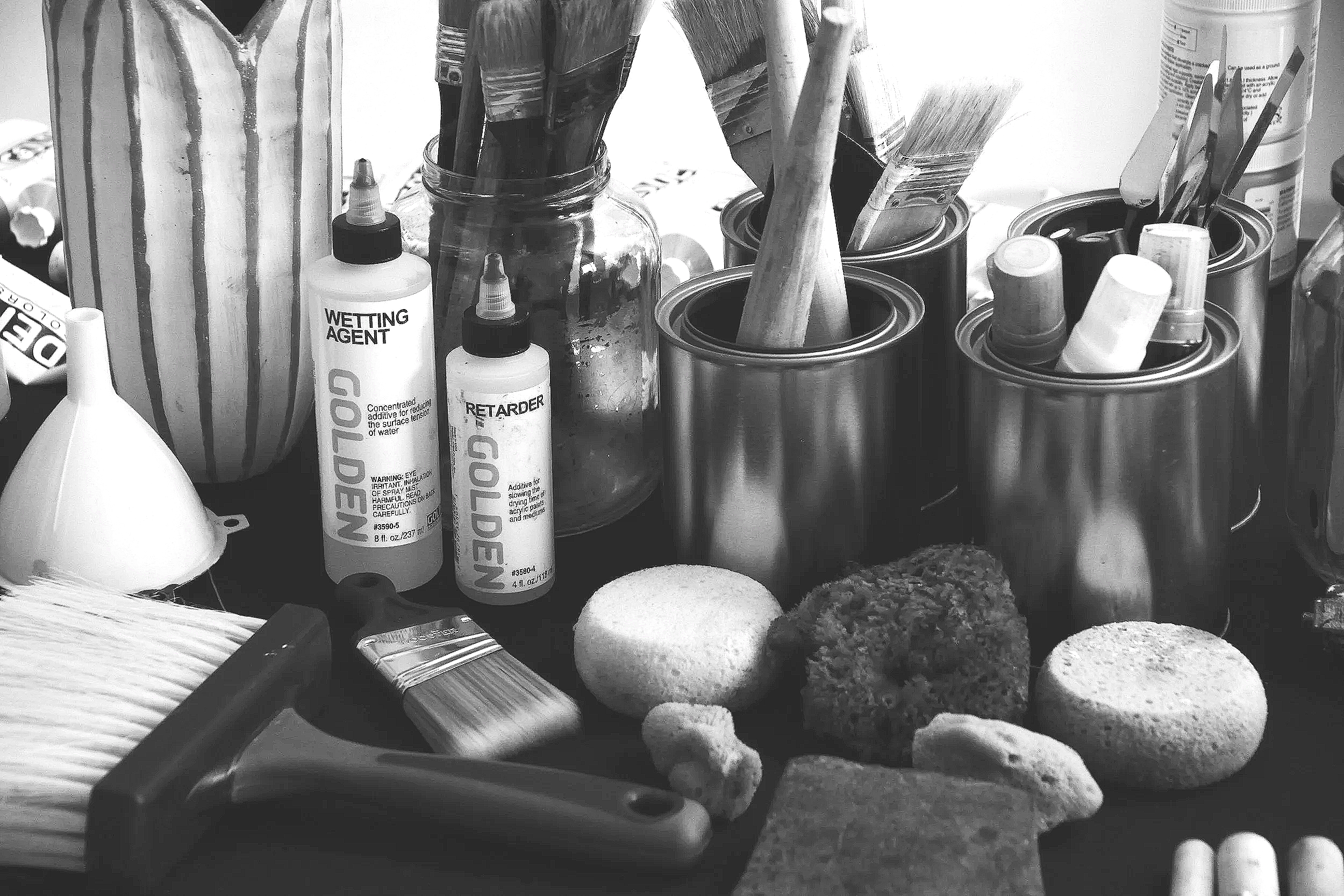 A black-and-white photograph documenting the painting tools and materials used by contemporary abstract artist Christopher Durst. Brushes, acrylic mediums, sponges, and mixed media supplies are arranged within the artist's Austin, Texas studio, offer