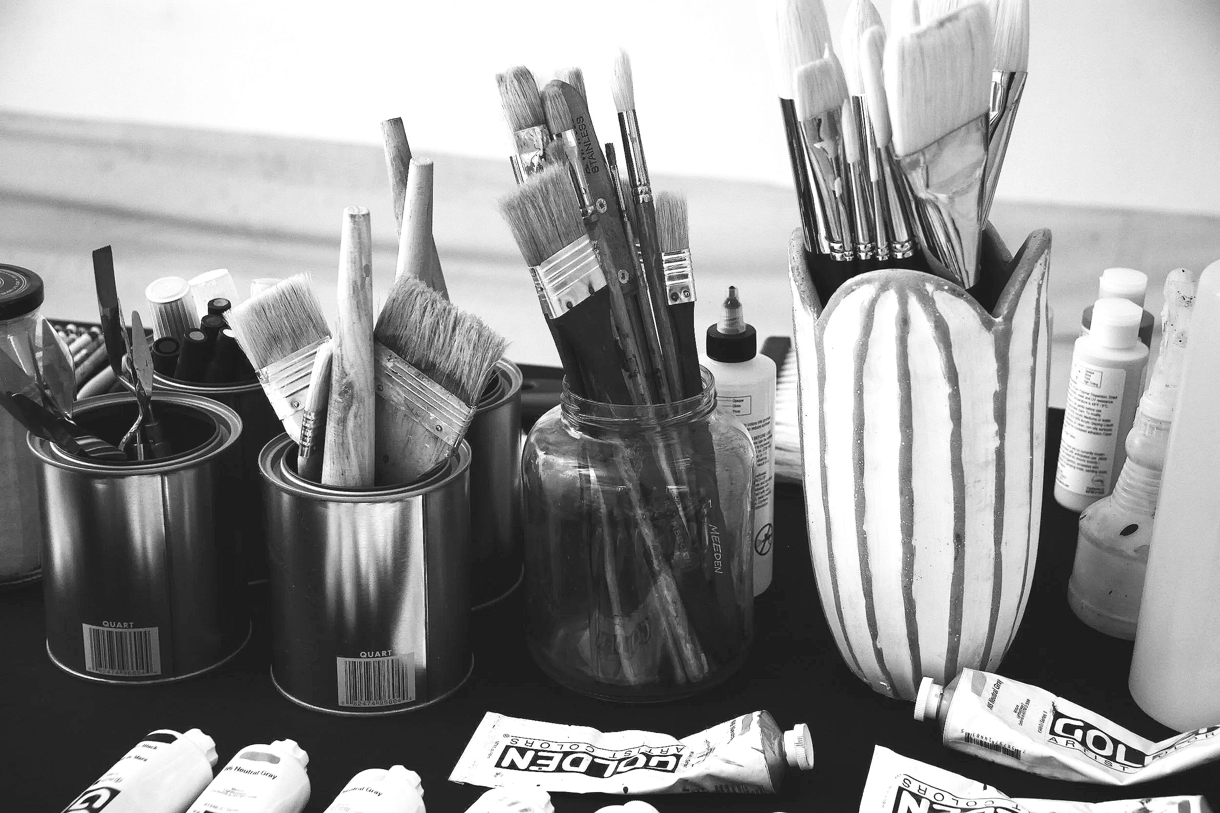 A black-and-white photograph of the personal studio of abstract artist Christopher Durst, featuring paintbrushes, acrylic paints, and working materials arranged across a studio table. The image offers a glimpse into the creative process behind his la